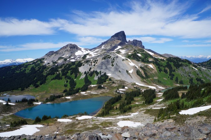 A photo of Black Tusk as seen from Panorama Ridge in Garibaldi Park on a blue sky day in the summer, with the alpine meadows and mountains in the background and lake in the foreground in BC.