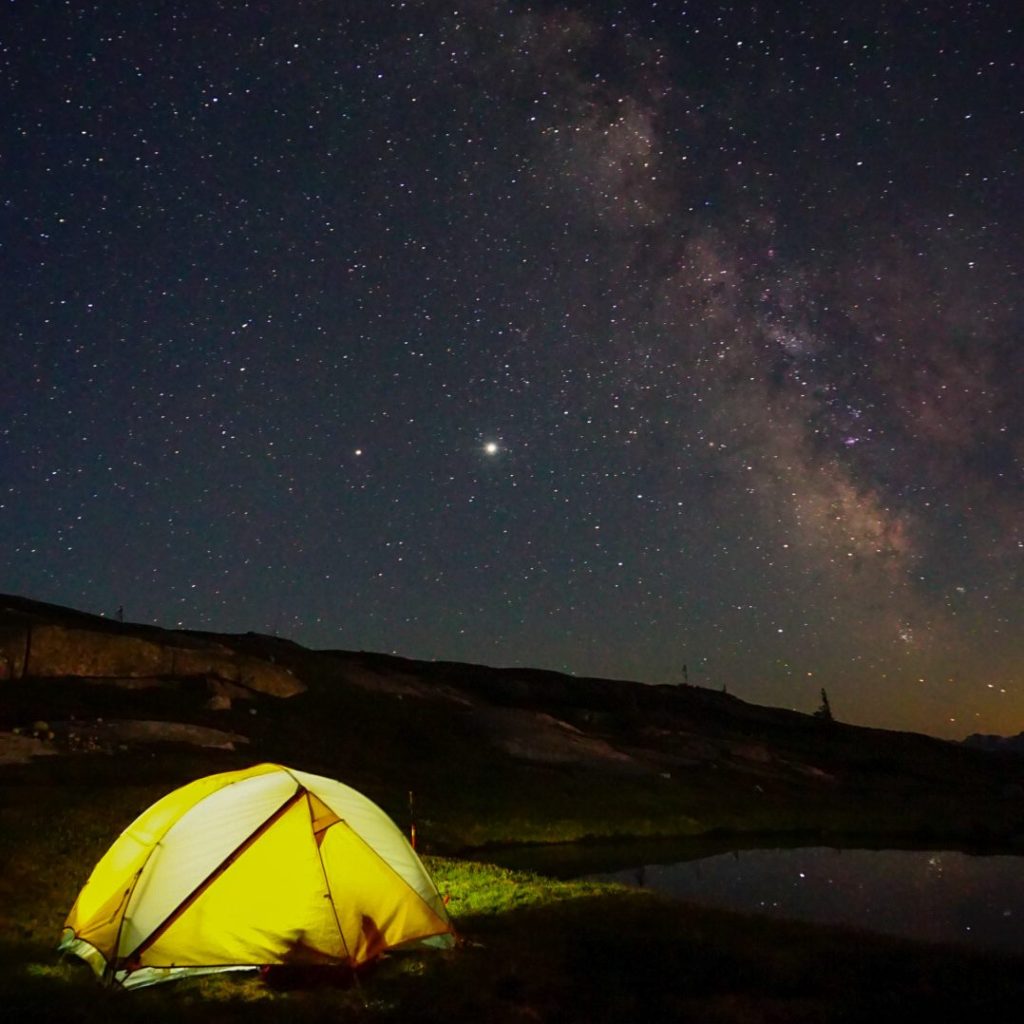 A yellow and grey north face tent is illuminated in the dark on a mountain peak with the milk way and starts in the background on top of Flatiron in Coquihalla Summit Rec Area near Hope, BC.