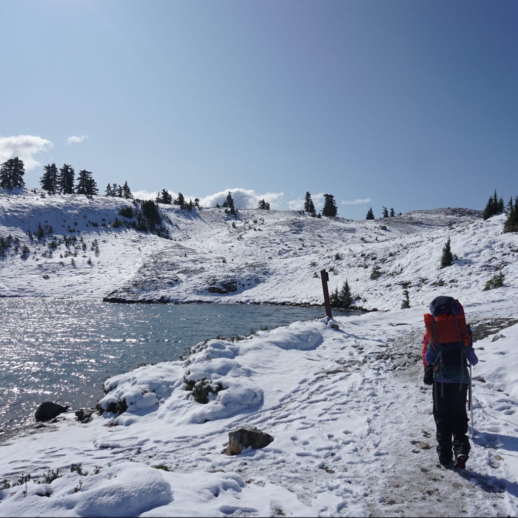 A girl guide walks next to Elfin Lakes with a large backpack in autumn when the ground is covered with a fresh layer of snow and beautiful sunny blue skies in Garibaldi Provincial Park in British Columbia