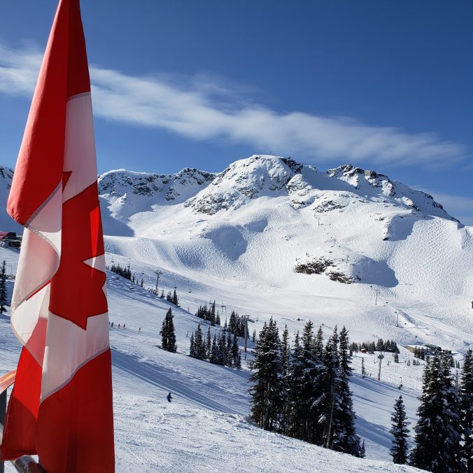 A photo of Peak Chair as seen from Roundhouse at Whistler. There is a Canadian flag in the foreground and snow covered mountain peaks on a blue sky day in the background, at Whistler, BC.