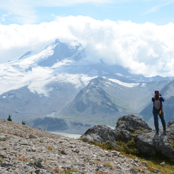 A female hiker stands back on to the camera on the top of Mount Price, staring at Mount Garibaldi, which is covered in glaciers and shrouded by clouds, in Garibaldi Provincial Park in British Columbia, Canada.