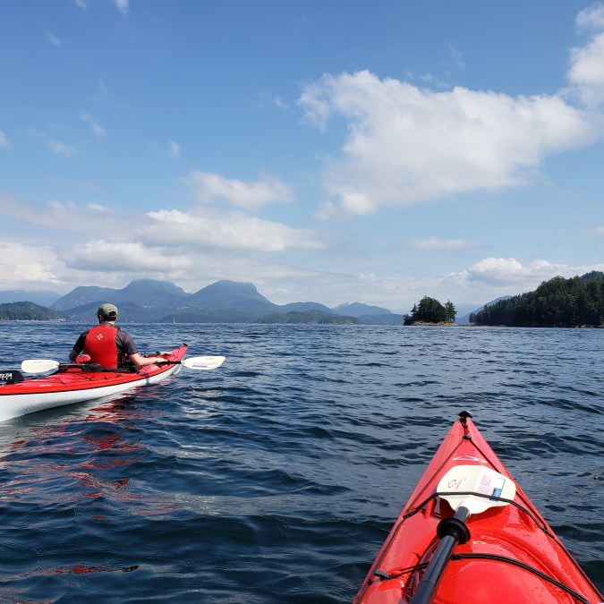 Two kayakers paddle on the Pacific Ocean towards Keats Island from Gibsons on the Sunshine Coast, BC on a sunny day, with the mountains in the background.