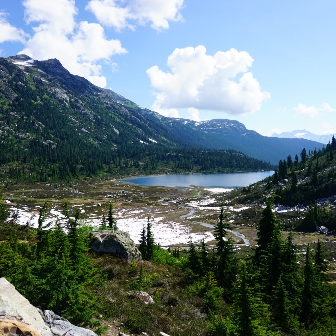 A photo of hanging lake from the ridgeline with snow still remaining in the meadow and the surrounding mountains, on the Rainbow Lake Trail in Whistler, British Columbia, Canada.