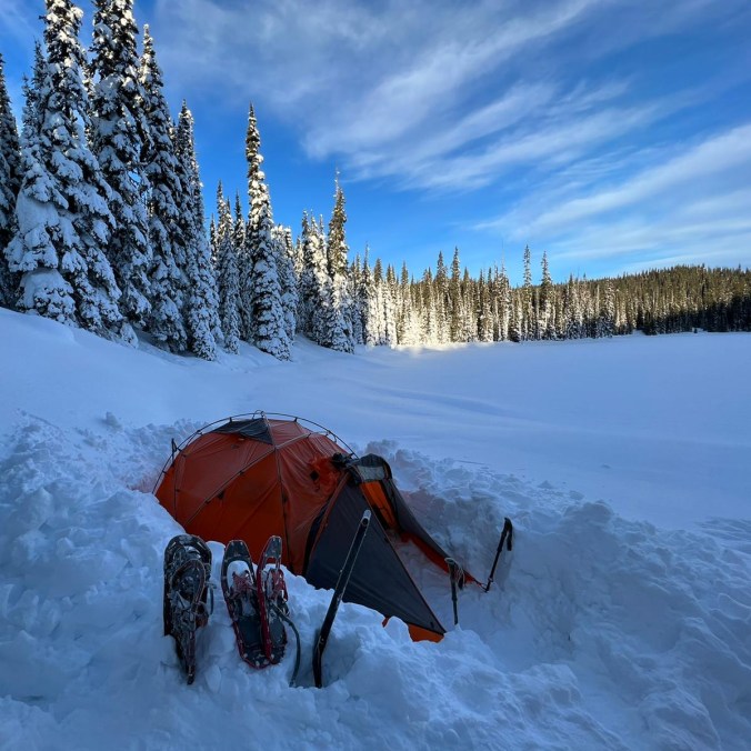 An orange tent is set up in the snow for camping with snowshoes and shovels outside the tent and a large snow covered lake and snowy trees in the background, on a sunny blue sky day at Poland Lake in E.C. Manning Provincial Park in British Columbia, Canada.