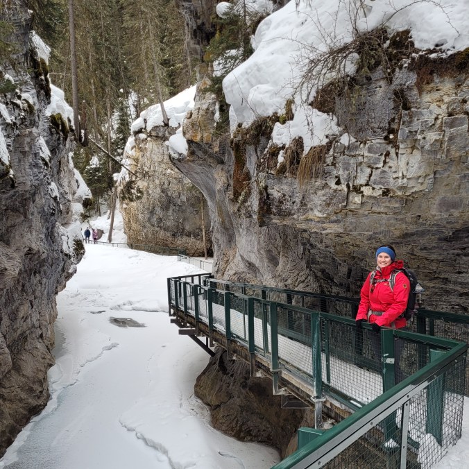 A winter female hiker stands on a metal walking bridge surrounded by a snow canyon with steep rock walls in Johnston Canyon in Banff National Park, in Alberta, Canada.
