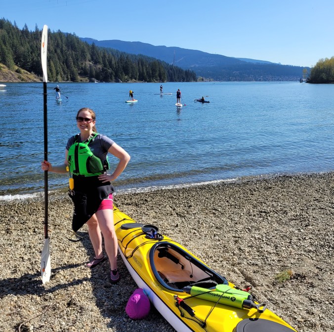A female kayaker wears a lifejacket and holds a paddle while smiling next to her yellow Boreal storm kayak on the rocky beach of Barnet Marine Park with the ocean and mountains in the background in Burnaby, British Columbia, Canada.