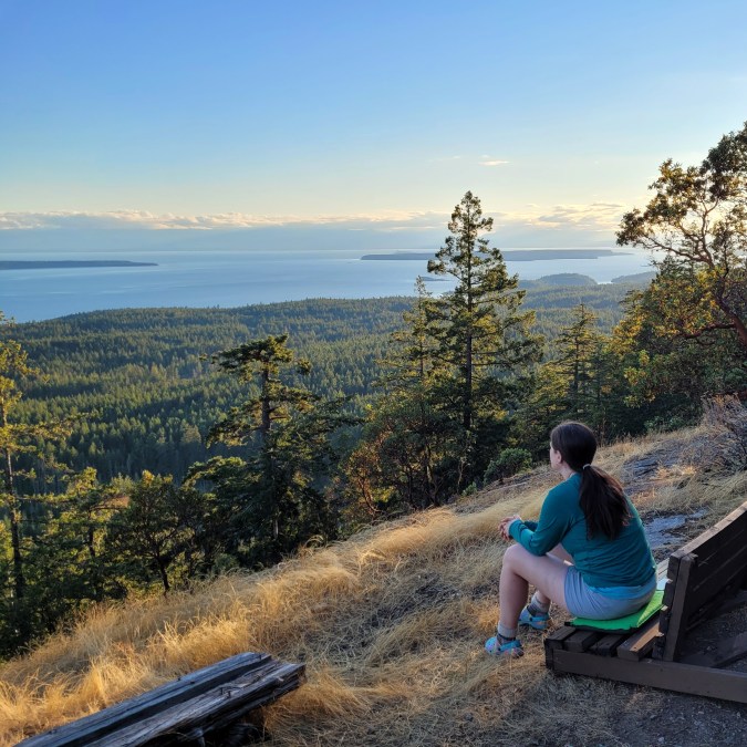 A female hiker / camper sits on a bench on Manzanita Bluff over looking the trees and blue water of the Salish Sea along the Sunshine Coast Trail in British Columbia, Canada.