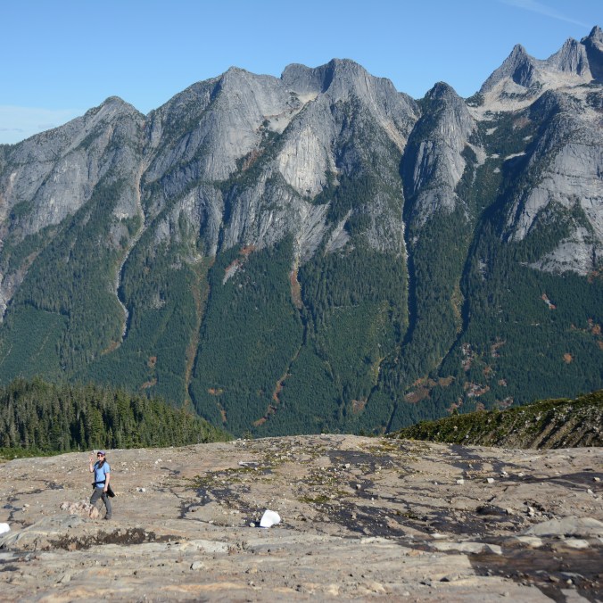 A small hiker and her dog pose on exposed rock with the large towering Slesse Mountains and forest behind them in the wilderness east of Chilliwack in British Columbia Canada on the Slesse Memorial trail.