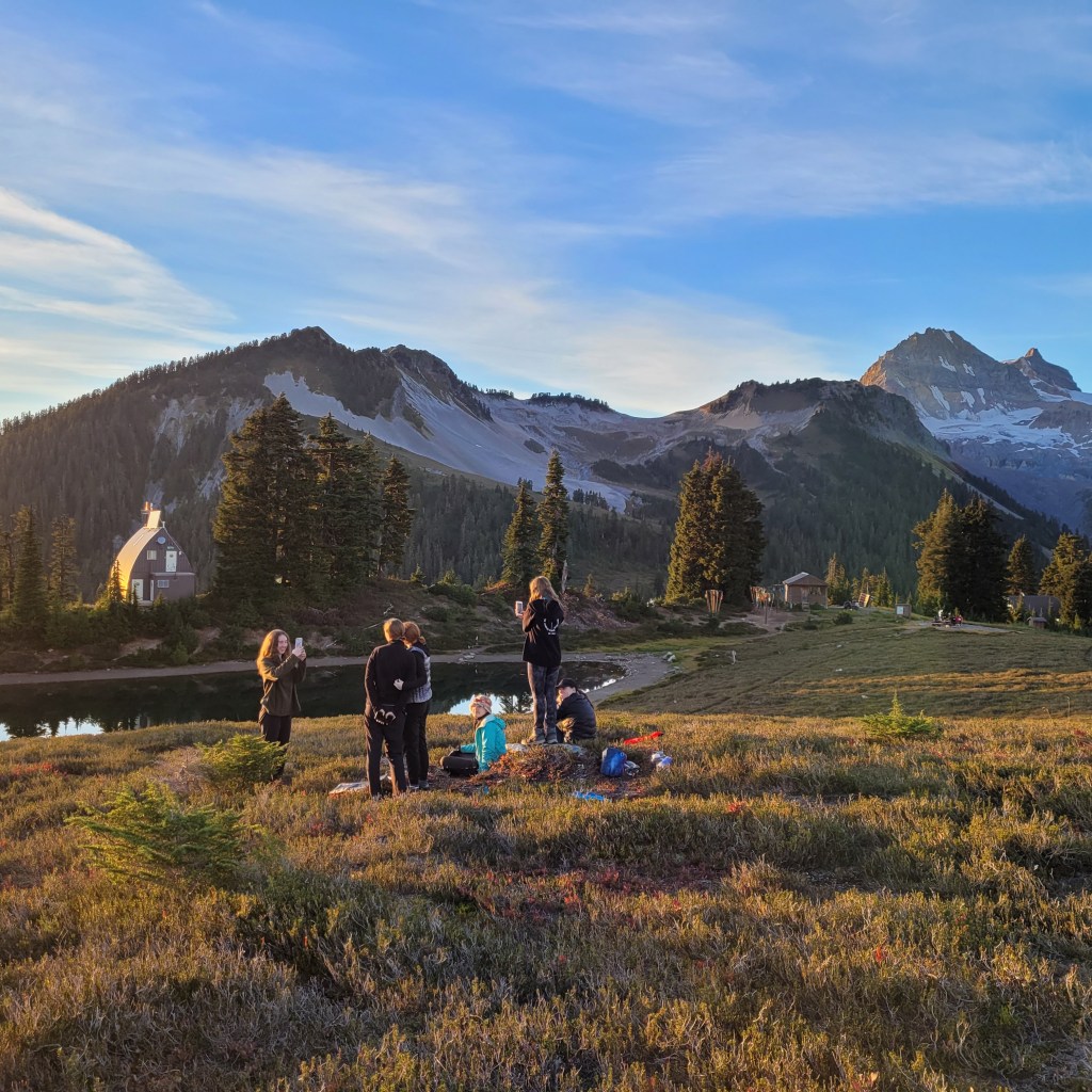 A girl guide group take photos of each other in front of Elfin Lakes, with the lake and the ranger hut in the background and the large mountain leading up to the saddle and Diamond Head in Garibaldi Provincial Park in British Columbia, Canada.