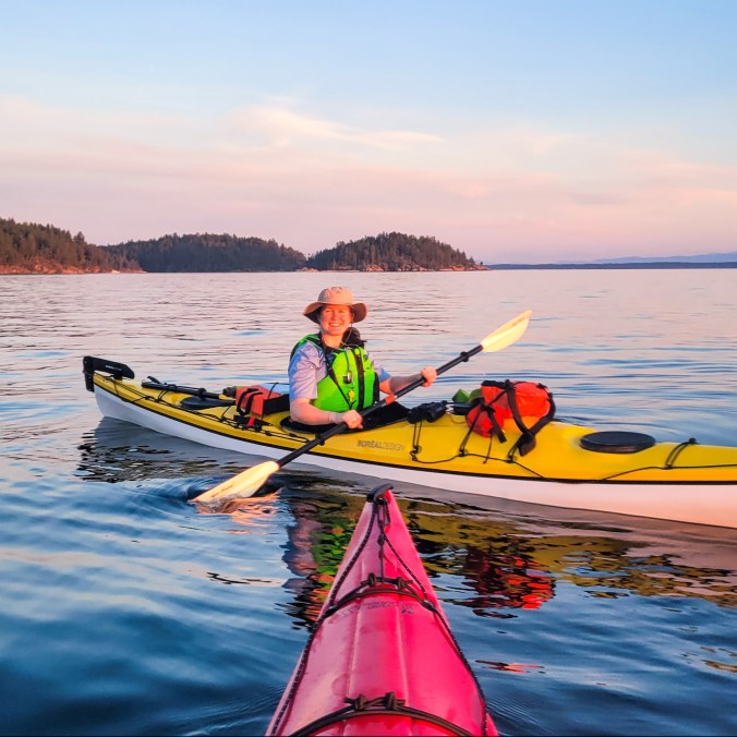 A female kayaker smiles at the camera from her yellow Boreal Storm kayak while the sun sets over the water and leaves a pink glow over everything in the Copeland Islands Provincial Park on the Sunshine Coast in British Columbia, Canada.