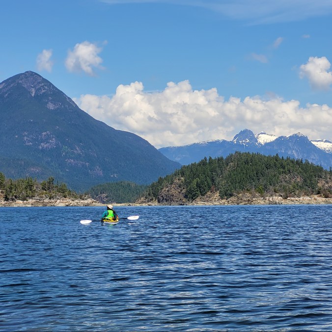 A female kayaker in a yellow kayak admires the view on the water with the mountains in the background in Desolation Sound Provincial Park on the Sunshine Coast in British Columbia, Canada.