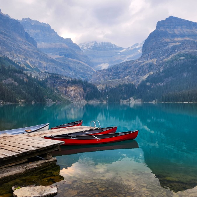 A photo of red canoes on the bright blue lake water of Lake O'Hara, with the mountains in the background on a cloudy day in Yoho National Park, in British Columbia, Canada.