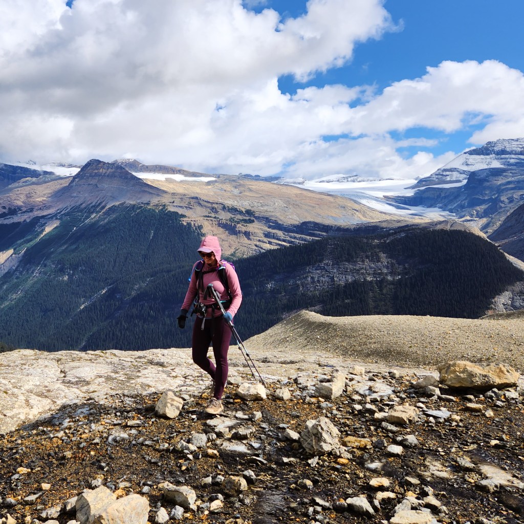 A female hiker walks with her hiking poles and hood up, facing away from the background view of large mountains and glaciers on the Iceline Trail in Yoho National Park in British Columbia, Canada.
