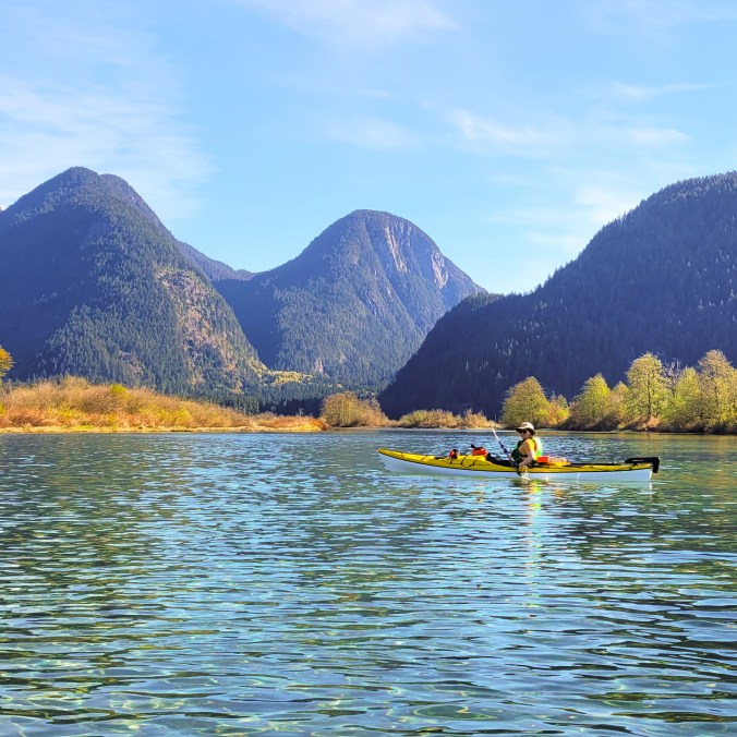 A woman in a yellow kayak paddles in crystal clear blue water on a sunny day with mountains in the background and a blue sky in Widgeon Creek by Widgeon Marsh in British Columbia, Canada.