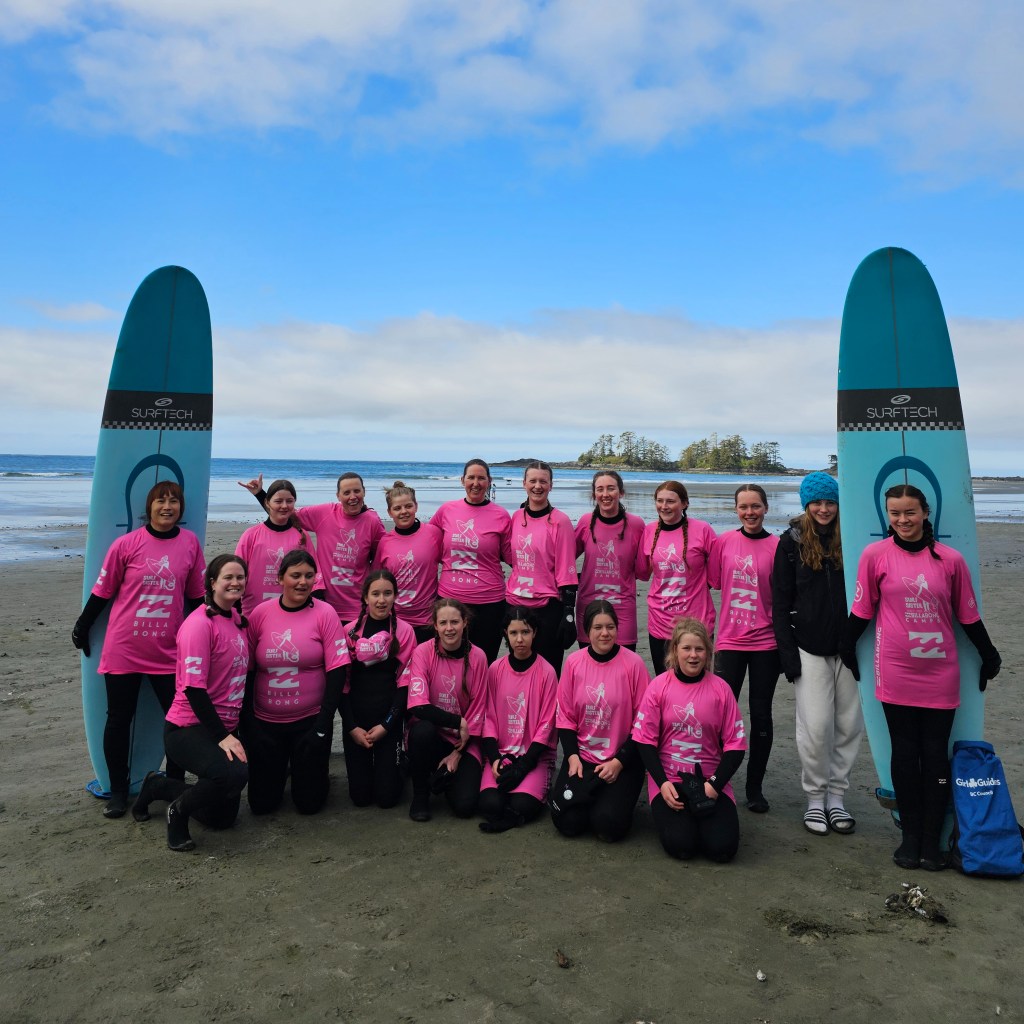 A group of female girl guides pose in pink shirts with 2 surf boards on the sandy beach in front of the Pacific Ocean at Chesterman Beach in Tofino on Vancouver Island, British Columbia.