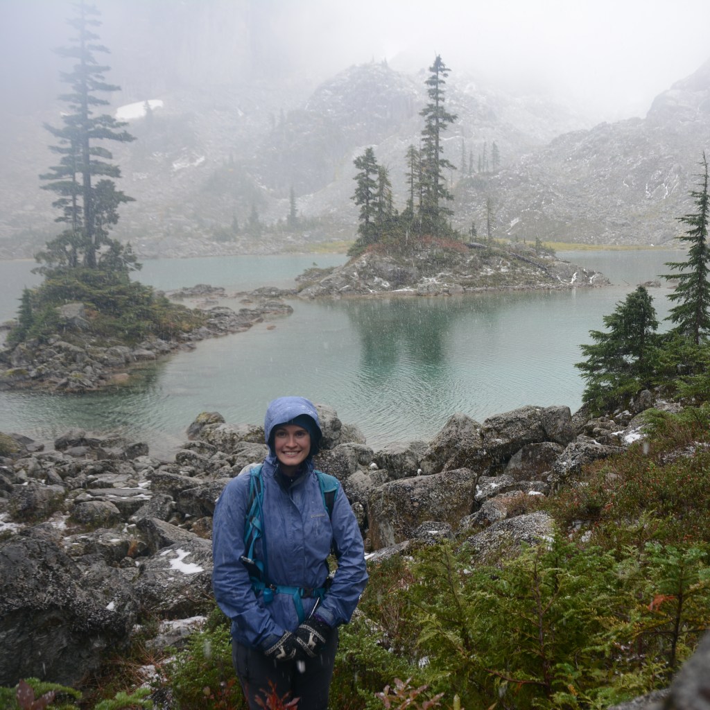 A female hiker looks at the camera on a very rainy day in front of Watersprite Lake in the alpine, outside of Squamish, BC.