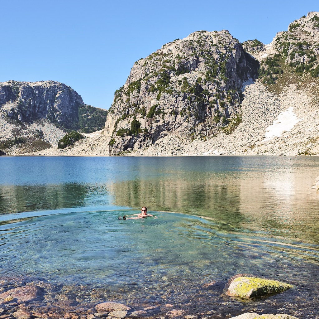 A female hiker floats in an alpine lake while swimming at Blanca Lake, with the mountains in the background on a blue sky, sunny day, north of Squamish, BC