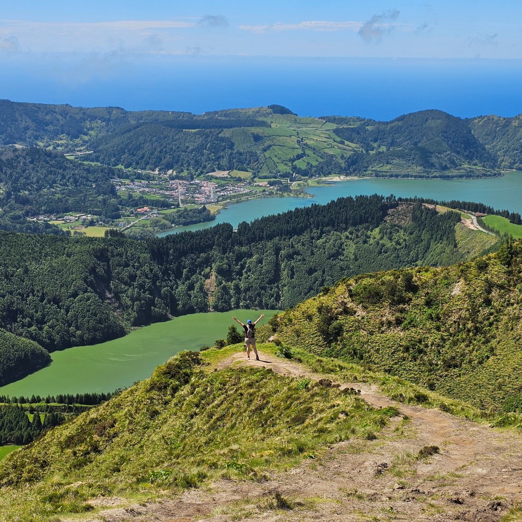 A female hiker stands on the edge of a cliff and raises her hands to the sky with the lakes, mountains, forest, and Atlantic ocean in the background at Sete Cidades on Sao Miguel Island in the Azores, Portugal.