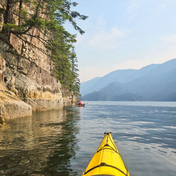 Two kayakers paddle up the still waters of Indian Arm, with the mountains in the background and a steep cliff extending into the water near Vancouver, BC.