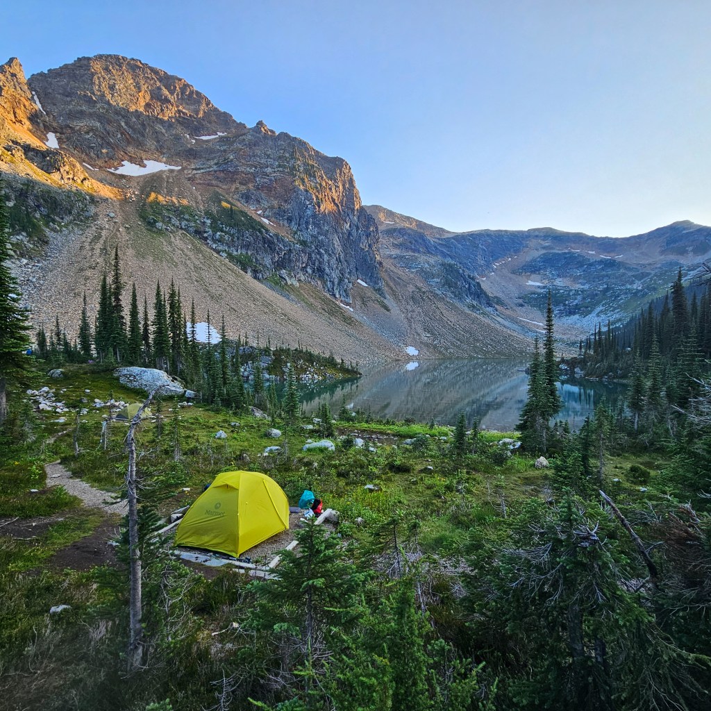 A photo of a tent in a meadow just before sunrise, as the sun turns the surrounding mountains orange on a clear sky day overlooking the alpine and forest around Jade Lake in Mount Revelstoke National Park in BC.