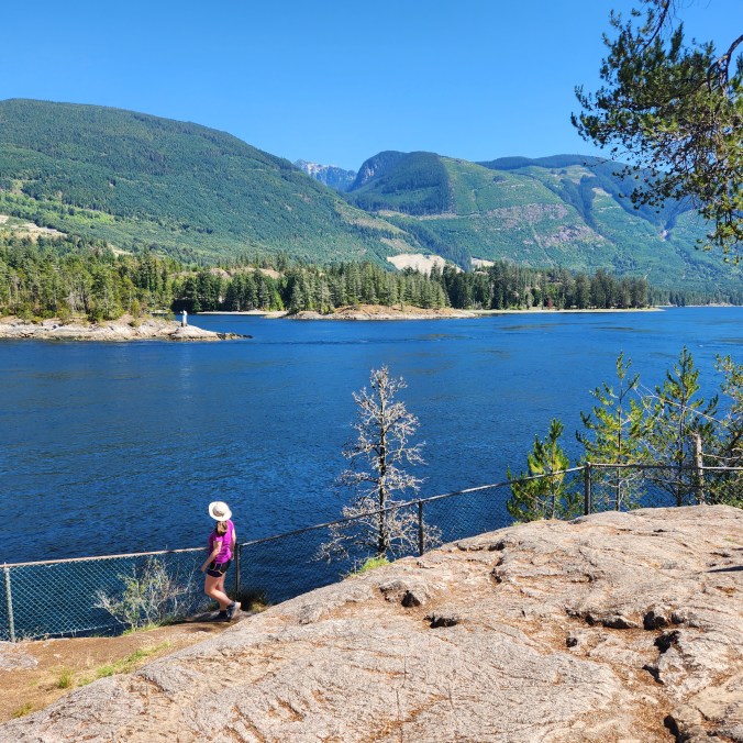 A female hiker stands next to a fence, looking out at the fast moving water of Sechelt Inlet near Skookumchuck Narrows, with the mountains in the background on a sunny, summer day, on the Sunshine Coast, BC.