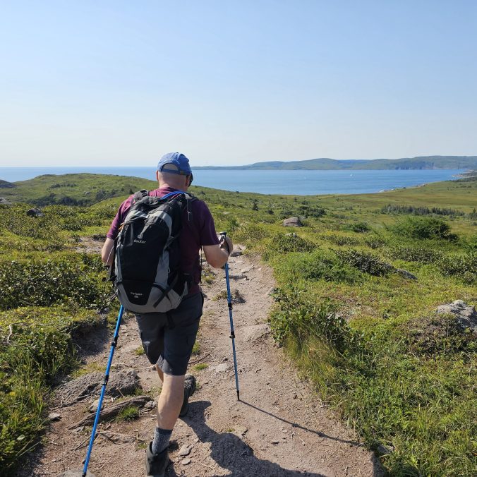 A hiker walks along a worn track on the East Coast Trail, Cape Spear Path section, with the ocean in the background on a sunny day in Newfoundland.