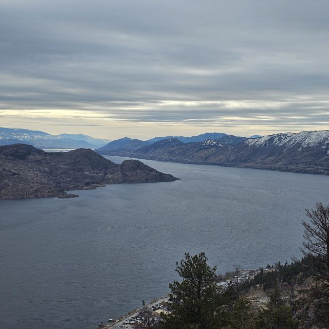 A photo of Okanagan Lake from the top of Pincushion Trail near Kelowna on a cloudy day with the mountains in the background in BC.