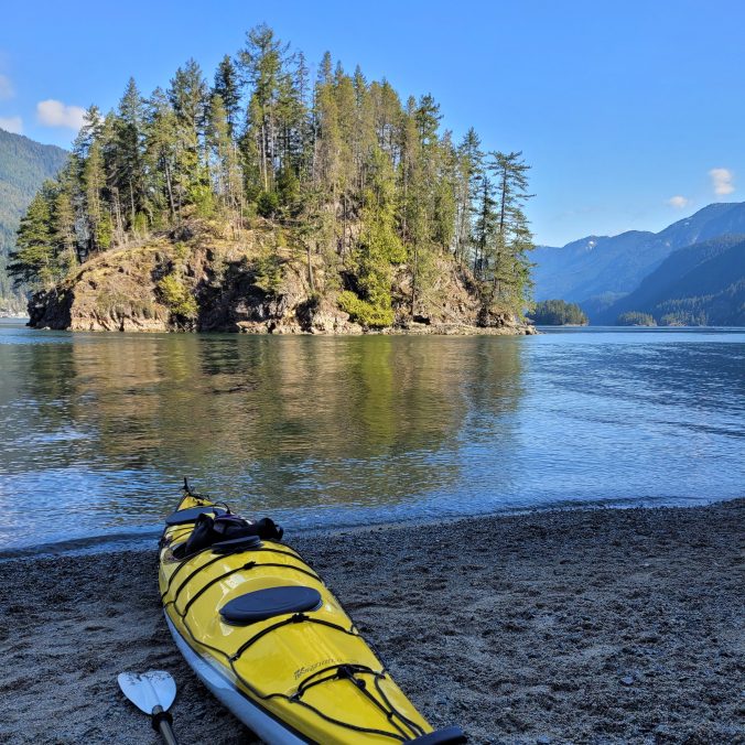 A photo of a yellow kayak resting on the beach in front of Jug Island on a sunny, blue sky day in Belcarra, BC.