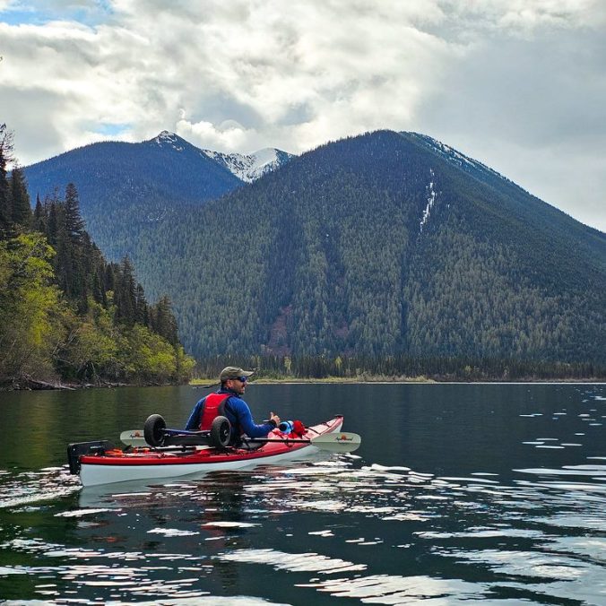 A kayaker paddles calm water in the morning on Isaac Lake on the Bowron Circuit, with mountains in the background in Northern BC.
