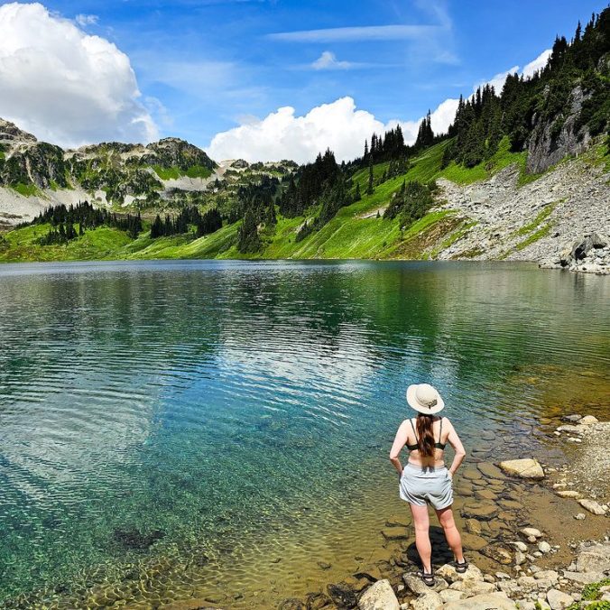 A female hiker stands back on in front of an alpine lake on a blue sky, sunny day, with the alpine meadows and mountains in the background, at Cirque Lake in the Callaghan Valley of Whistler, BC.