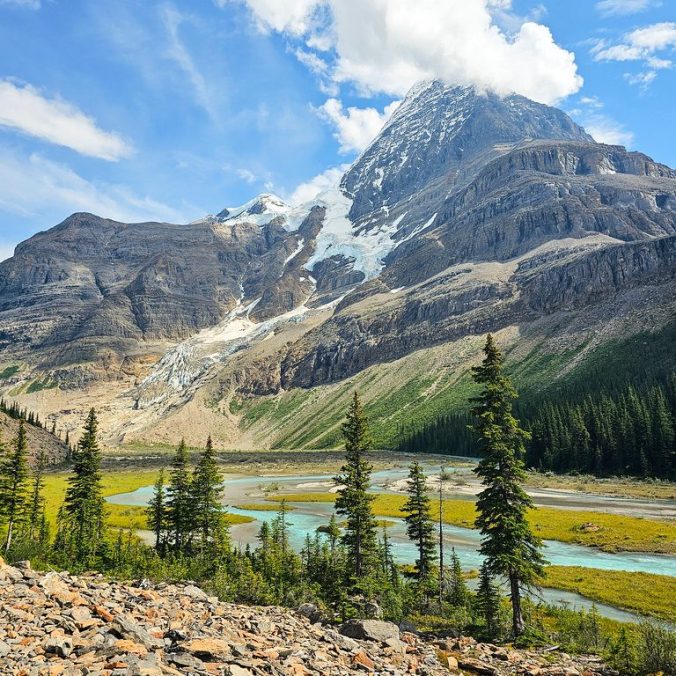 A photo of Mount Robson on a sunny day with the Robson River in and trees in the foreground and the mountains and glacier in the background, on the Berg Lake Trail in Mount Robson Provincial Park, BC.