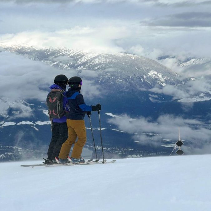 Two female skiers on the top of Revelstoke Ski Resort, with the mountains and slopes in the background on a cloudy day in British Columbia.