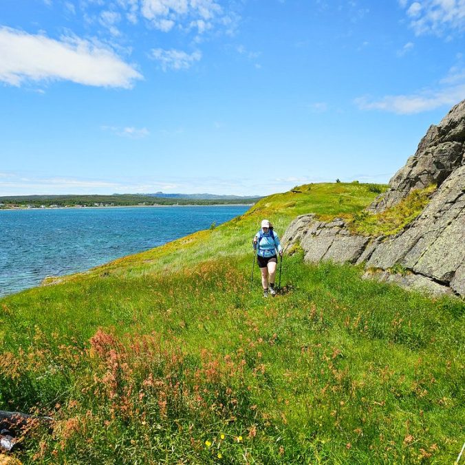 A female hiker walks across a grassy meadow with the atlantic ocean in the background on the Bear Cove Point Trail on the East Coast Trail in Newfoundland.