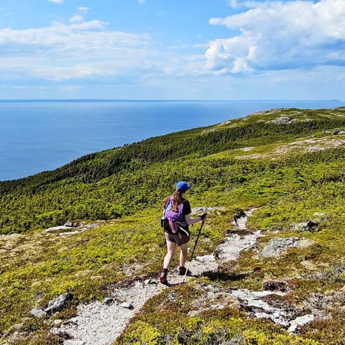 A female hiker walks along a gravel path trail on a grassy bluff overlooking the Atlantic Ocean on White Horse Path of the East Coast Trail in Newfoundland.