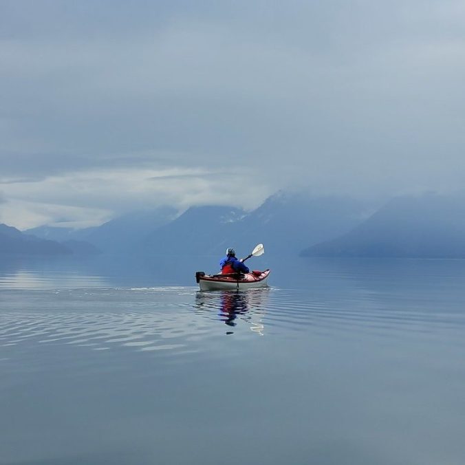 A female kayaker paddles on Harrison Lake on a cloudy morning with flat water and the mountains in the background.