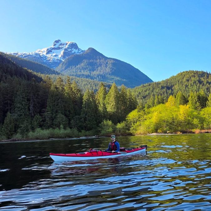 A kayaker paddles up Pitt Lake with Raven Creek Campsite and Golden Ears Peak in the background on a sunny day in Pitt Meadows, BC.