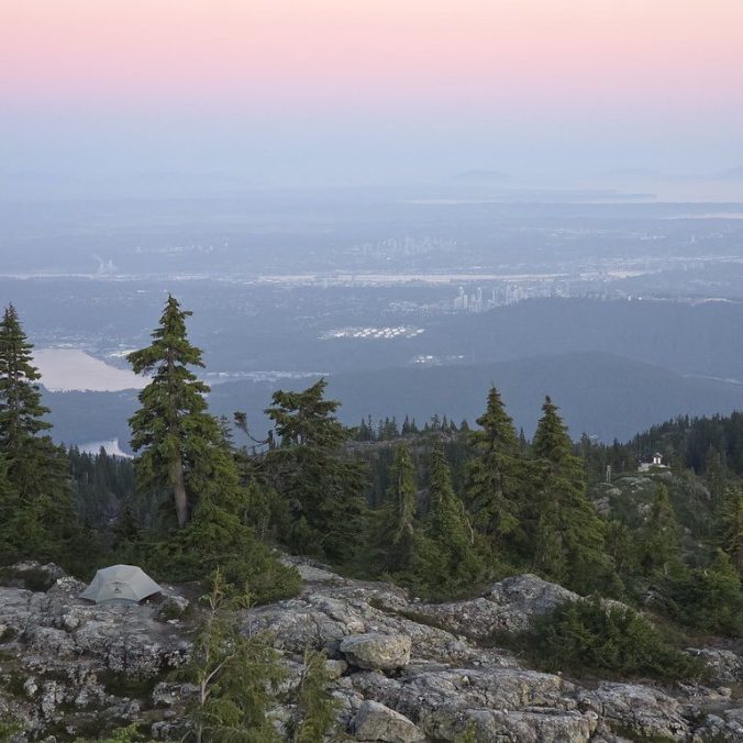 A photo of the sunset over Vancouver City as seen from Pump Peak in Mount Seymour Provincial Park, with a tent in the foreground and the pink hues of the sunset and the city in the background.
