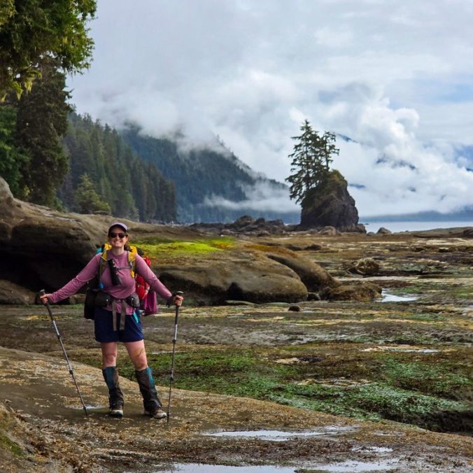 A female backpacker smiles at the camera on a rocky ocean shelf with a sea stack and the pacific ocean in the background on a cloudy day on the West Coast Trail on Vancouver Island, BC at Owen Point.