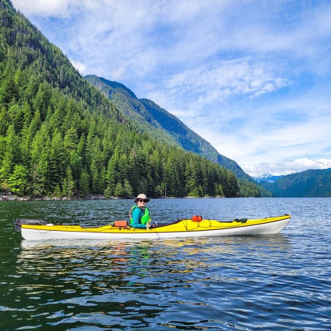 A female kayaker paddles Alouette Lake with the mountains in the background on a sunny summer day in Golden Ears Park, BC.