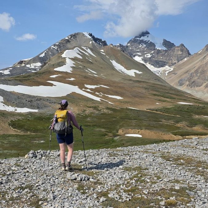 A female hiker stands back on to the camera with the Rocky Mountains in the background on a blue sky day at Parker Ridge in Banff National Park.