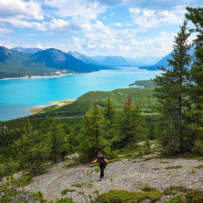 A female hiker climbs up a steep slope with the forest and mountains in the background and Lake Abraham on the Vision Quest Trail in Nordegg, Alberta.