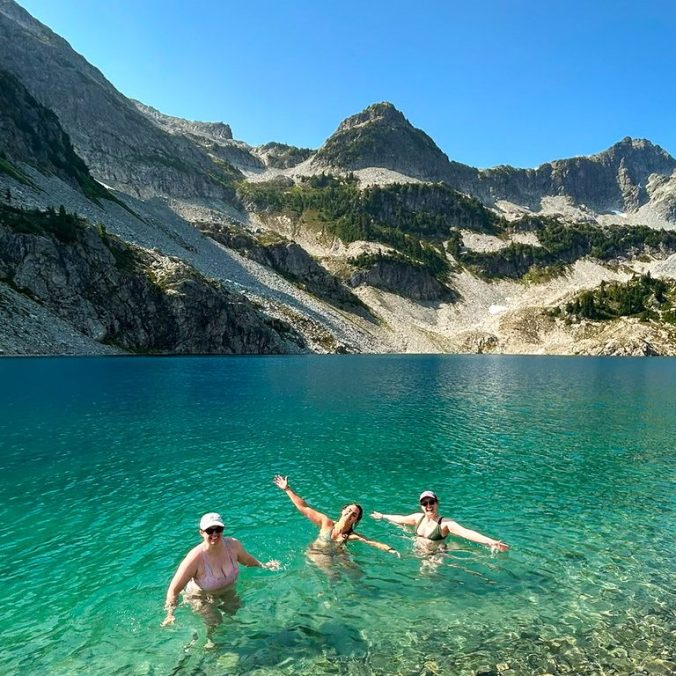 A group of three women swim in the bright blue waters of Tricouni Berg Lake with Tricouni Peak in the background on a sunny day outside Squamish, BC.